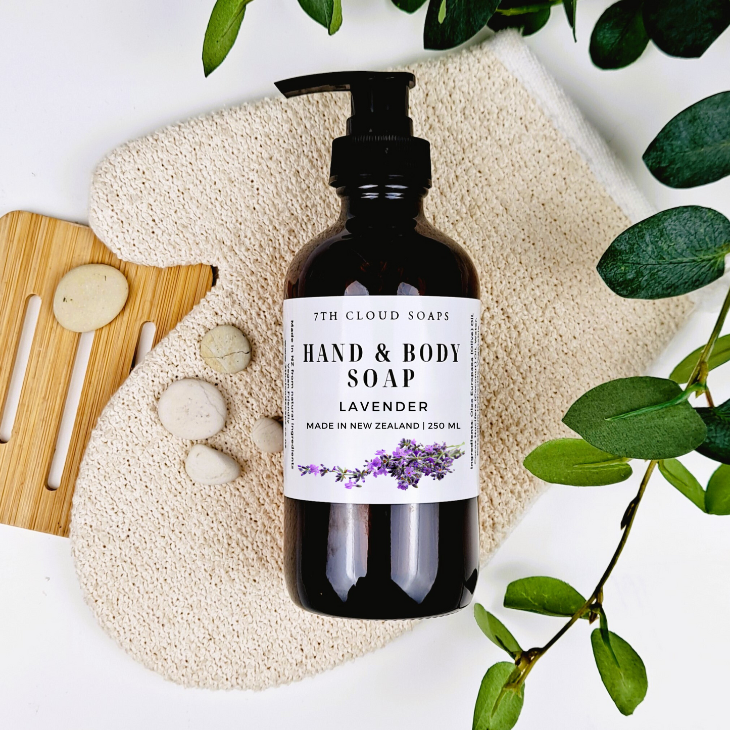 A 250ml glass bottle of liquid hand & body soap labeled 'Lavender', with a pump dispenser, alongside a natural loofah, massage stones, a bamboo brush, and green leaves.