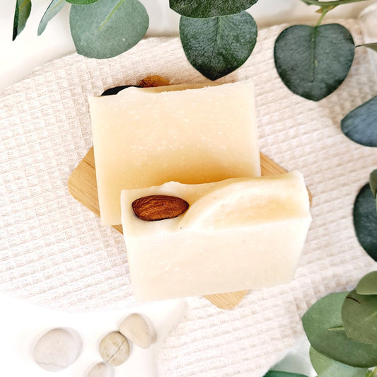 Two bars of soap with a small almond on a wooden board, surrounded by green leaves on a textured white surface.