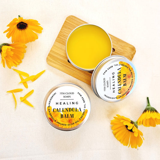 An open aluminium tin of Calendula Healing Balm with a yellowish substance inside, accompanied by whole calendula flowers and petals on a wooden surface.