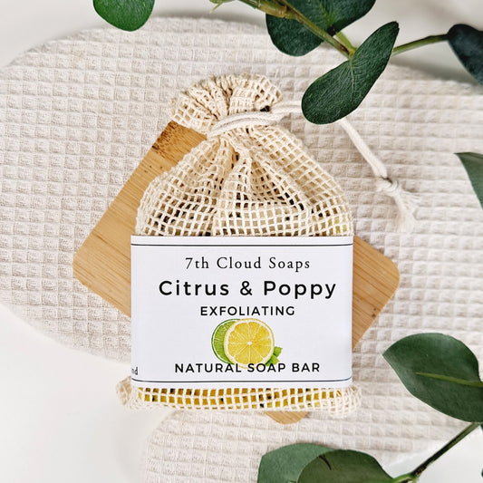 A natural soap bar labeled 'Citrus & Poppy Exfoliating' from 7th Cloud Soaps, packaged in a beige mesh net with a paper label, on a background with green leaves.