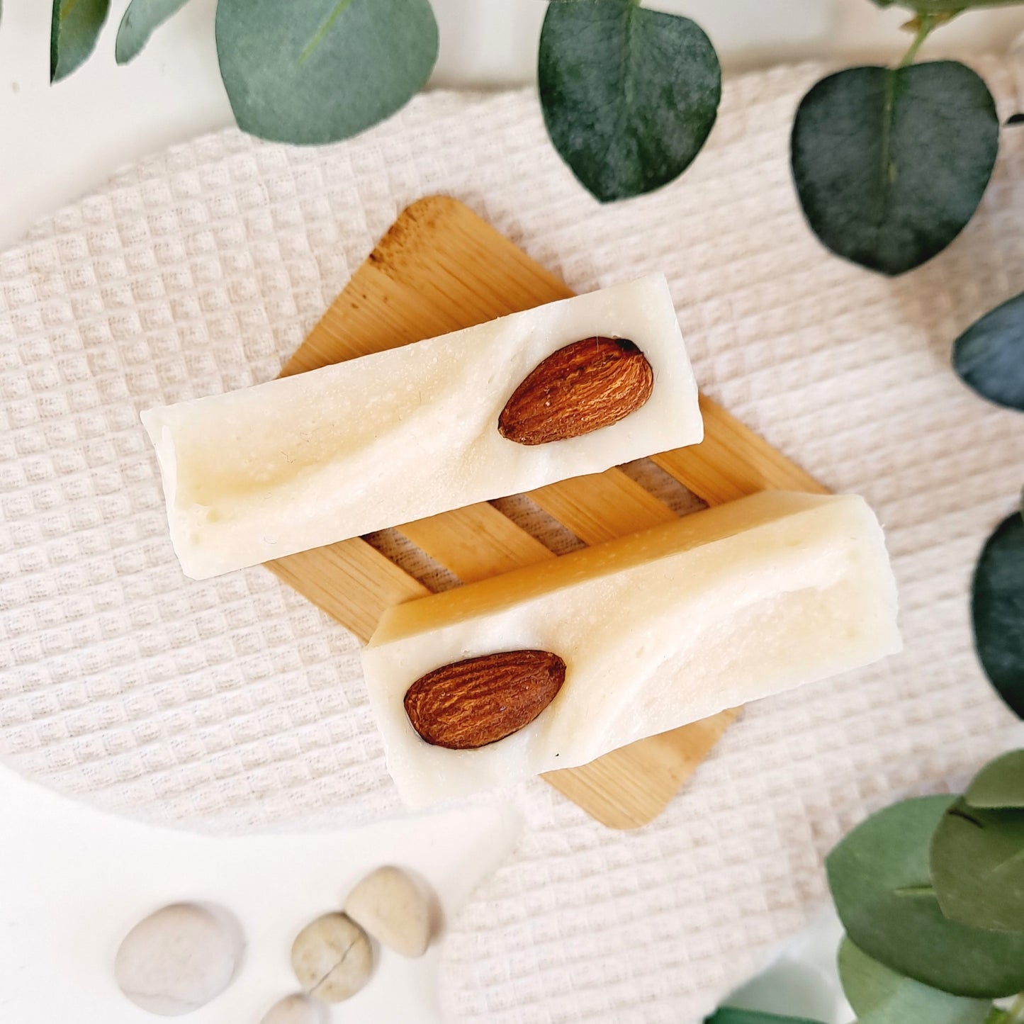 Wooden soap dish with soap bars and almonds on a textured surface with green leaves.