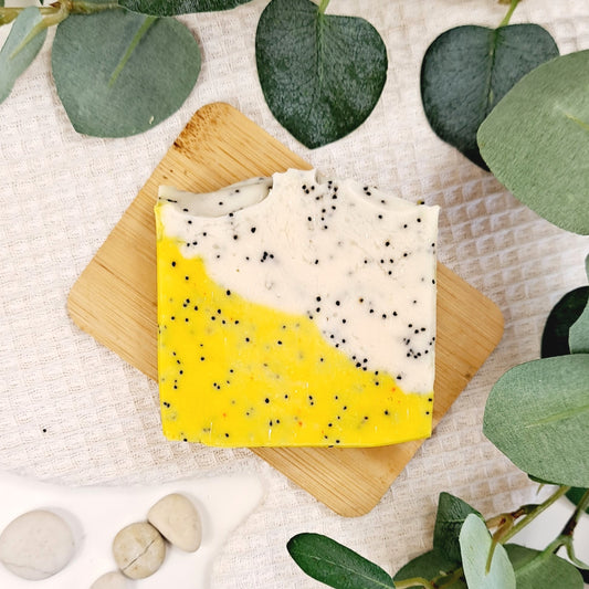 Two soap bars with yellow and white speckled design on a wooden board, surrounded by green leaves and stones.
