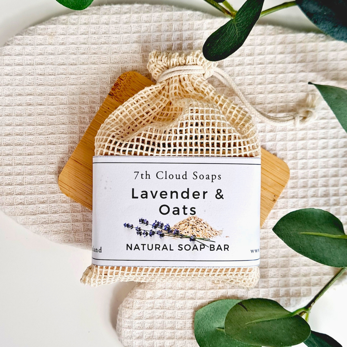 7th Cloud Soaps Lavender & Oats natural soap bar with a mesh bag on a textured surface with green leaves.