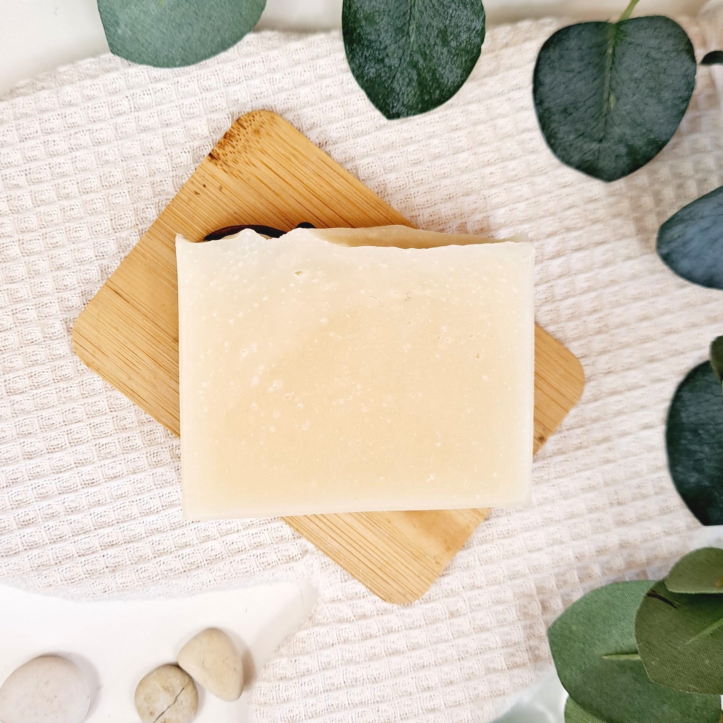 Bar of soap on a wooden soap dish with green leaves and stones on a textured white surface