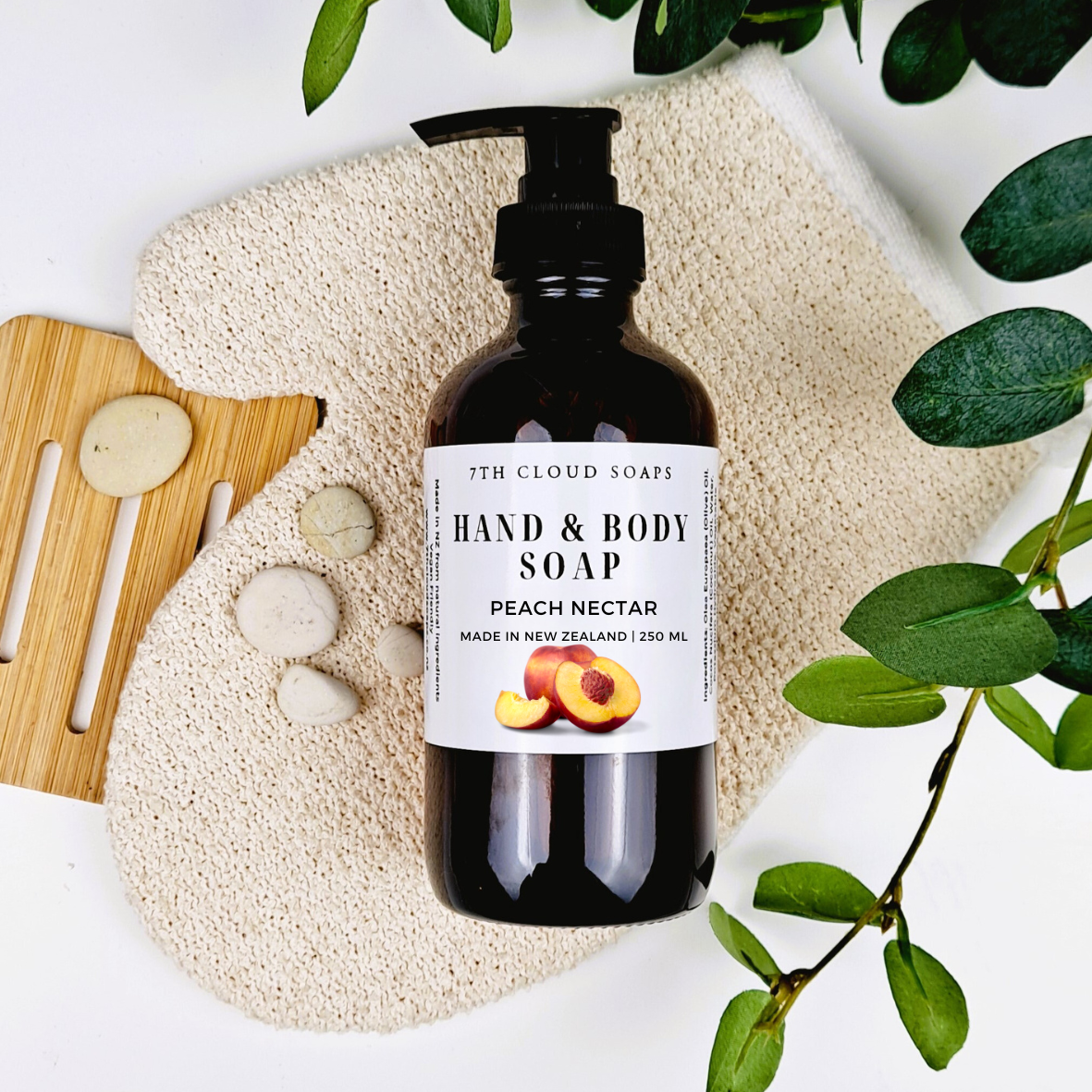 A 250ml glass bottle of liquid hand & body soap labeled 'Peach Nectar' from 7th Cloud Soaps, displayed with a pump, surrounded by a beige loofah, wooden soap dish