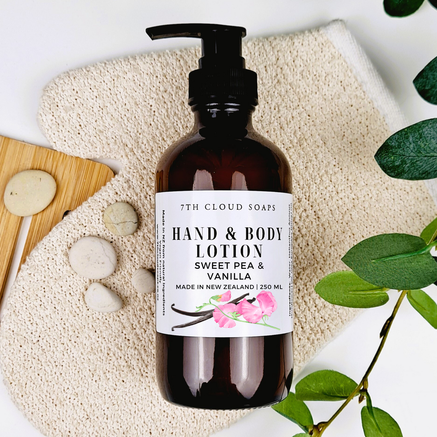 A pump bottle of hand & body lotion with the label 'Sweet Pea & Vanilla'. The bottle is placed on a natural-colored loofah, with a wooden comb, white soap tablets, and green leaves in the background.