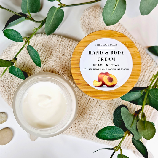 Hand and body cream jar with a wooden lid labeled 'Peach Nectar' on a textured surface with green leaves.