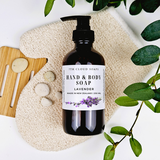 A 250ml glass bottle of liquid hand & body soap labeled 'Lavender', with a pump dispenser, alongside a natural loofah, massage stones, a bamboo brush, and green leaves.