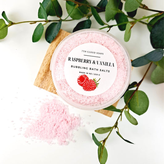 A container of Bubbling Magnesium Bath Salts with Raspberry & Vanilla scent, surrounded by green leaves. The container is labeled with the product name and the brand, 7th Cloud Soaps.
