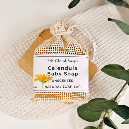 A bar of unscented natural baby soap with calendula, packaged in a net bag with a label that reads '7th Cloud Soaps, Calendula Baby Soap, Unscented, Natural Soap Bar'.