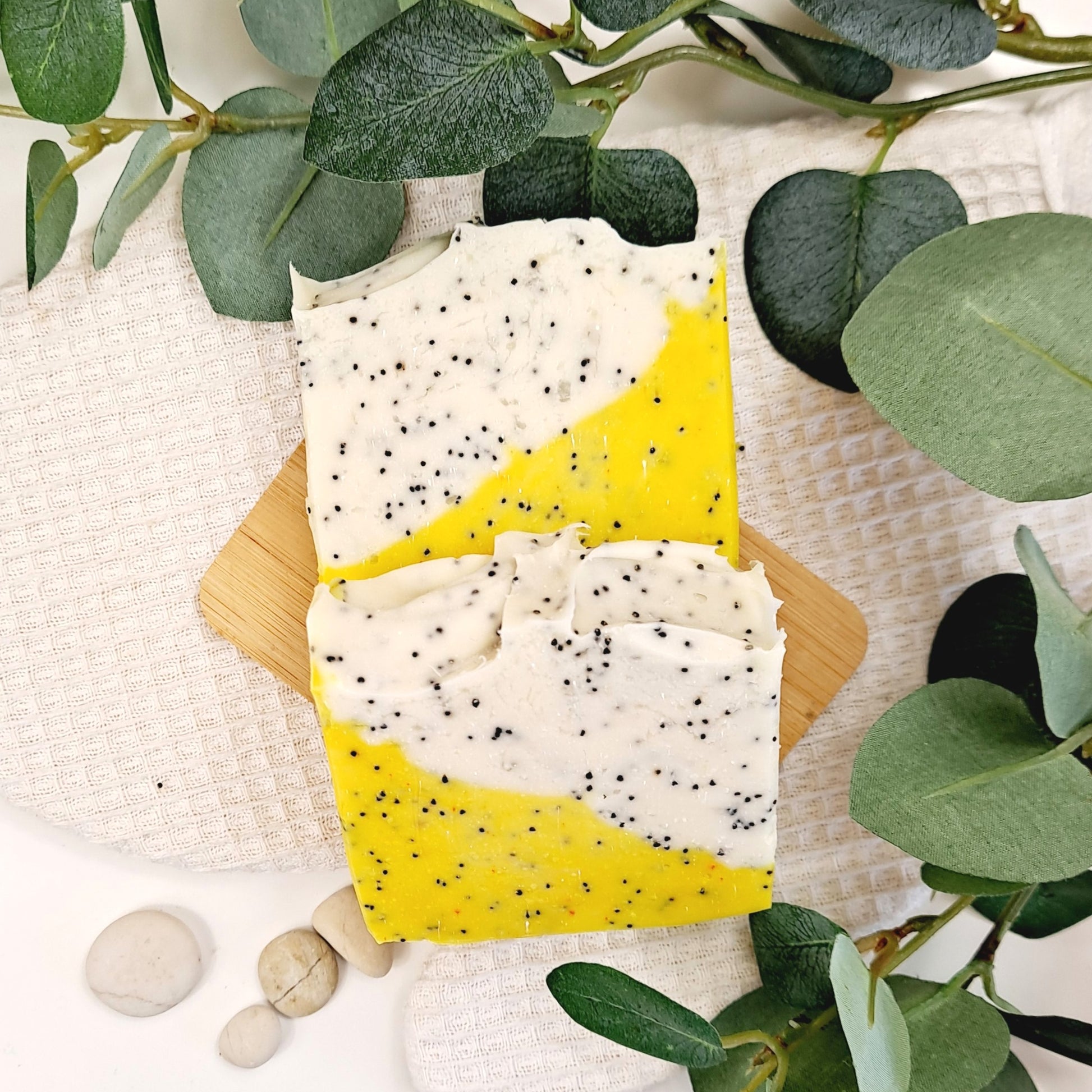 Two soap bars with a yellow and white gradient on a wooden board, surrounded by green leaves.