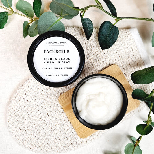 A jar of face scrub with jojoba beads and kaolin clay is open, revealing a light-colored product. It is placed on a wooden surface with a white background and green leaves around it.