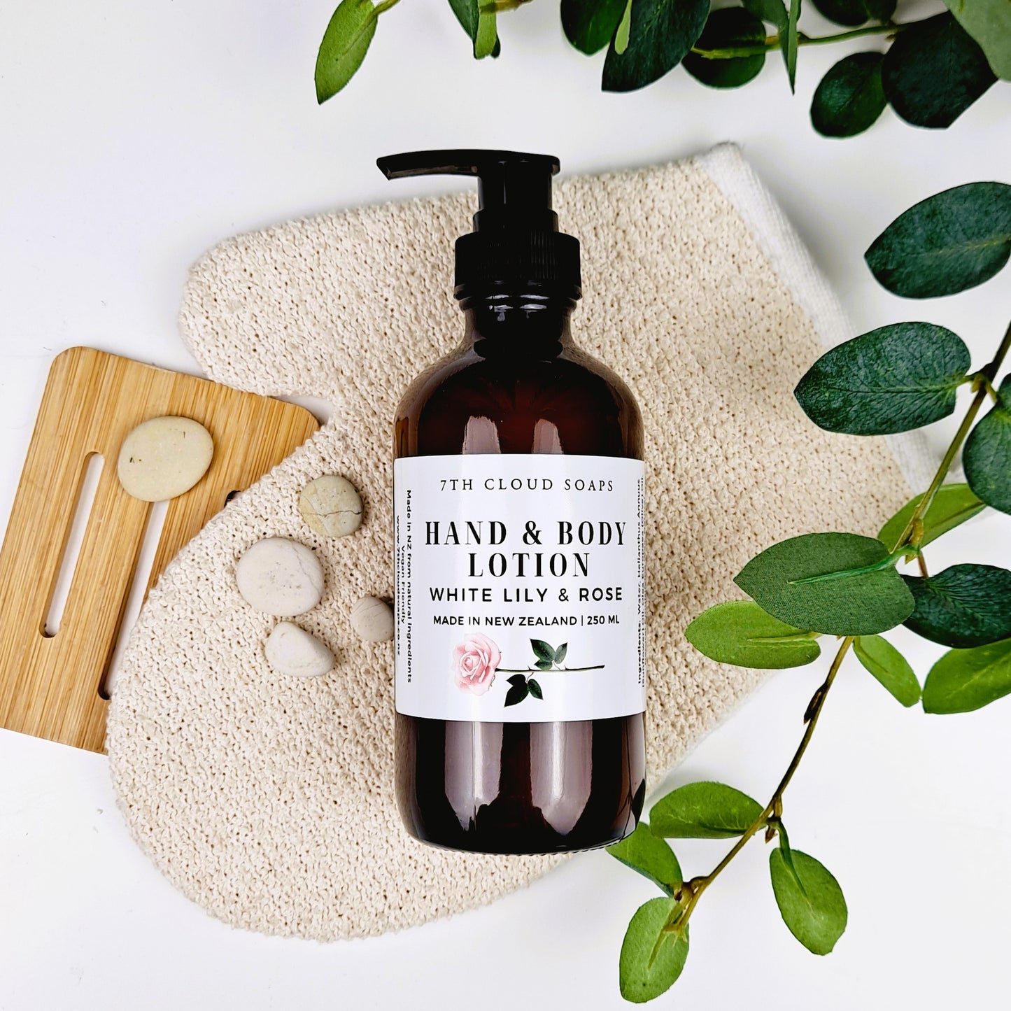 A pump bottle of Hand & Body Lotion - White Lily & Rose placed on a white towel with a wooden board, surrounded by green leaves and a white marble coaster.