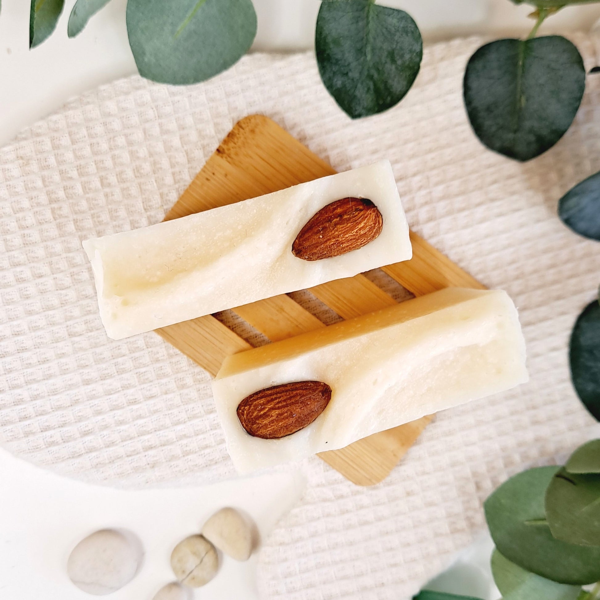 Wooden soap dish with soap bars and almonds on a textured surface with green leaves.