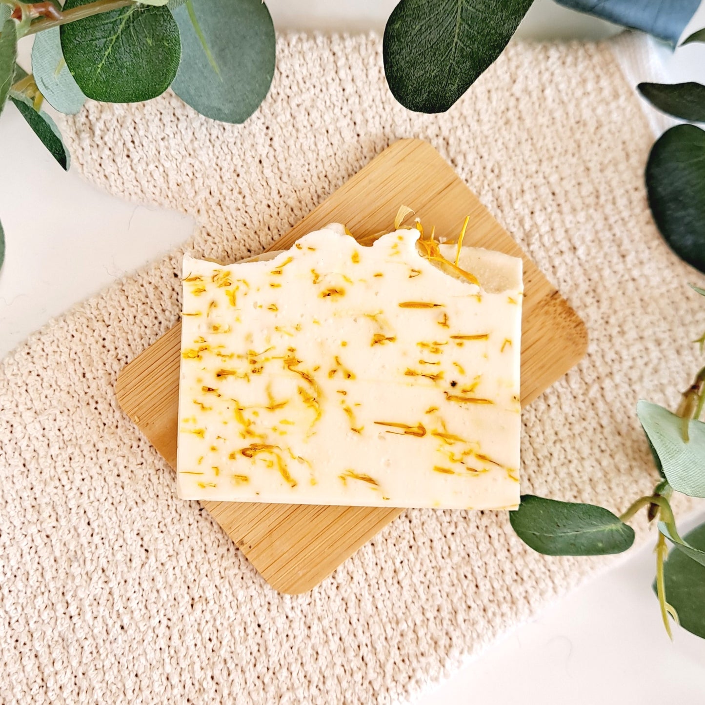 A natural soap bar labeled 'Calendula' from 7th Cloud Soaps, presented on a wooden soap dish, with a net bag, in a bathroom setting.