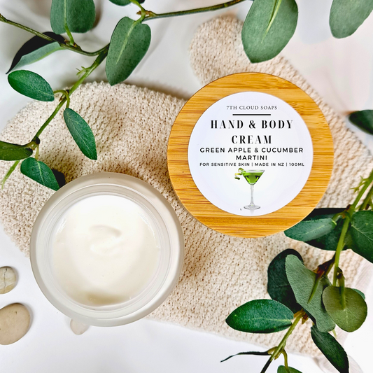 A glass jar with a bamboo lid labeled 'HAND & BODY CREAM' with a picture of a martini glass, placed on a towel surrounded by green leaves.