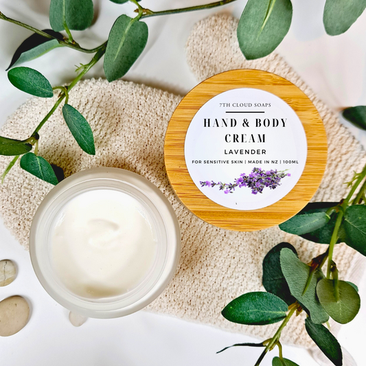 A glass jar with a bamboo lid labeled 'HAND & BODY CREAM' with a small amount of cream scooped out and displayed in front of the jar. The jar is placed on a beige textile with green leaves surrounding it.
