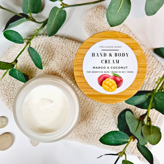 A glass jar with a bamboo lid labeled 'Hand & Body Cream, Mango & Coconut' placed on a natural colored loofah, surrounded by green leaves and a white stone.