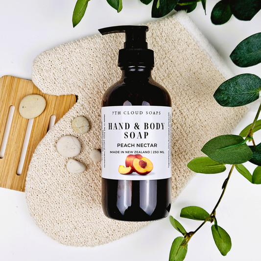 A 250ml glass bottle of liquid hand & body soap labeled 'Peach Nectar' from 7th Cloud Soaps, displayed with a pump, surrounded by a beige loofah, wooden soap dish