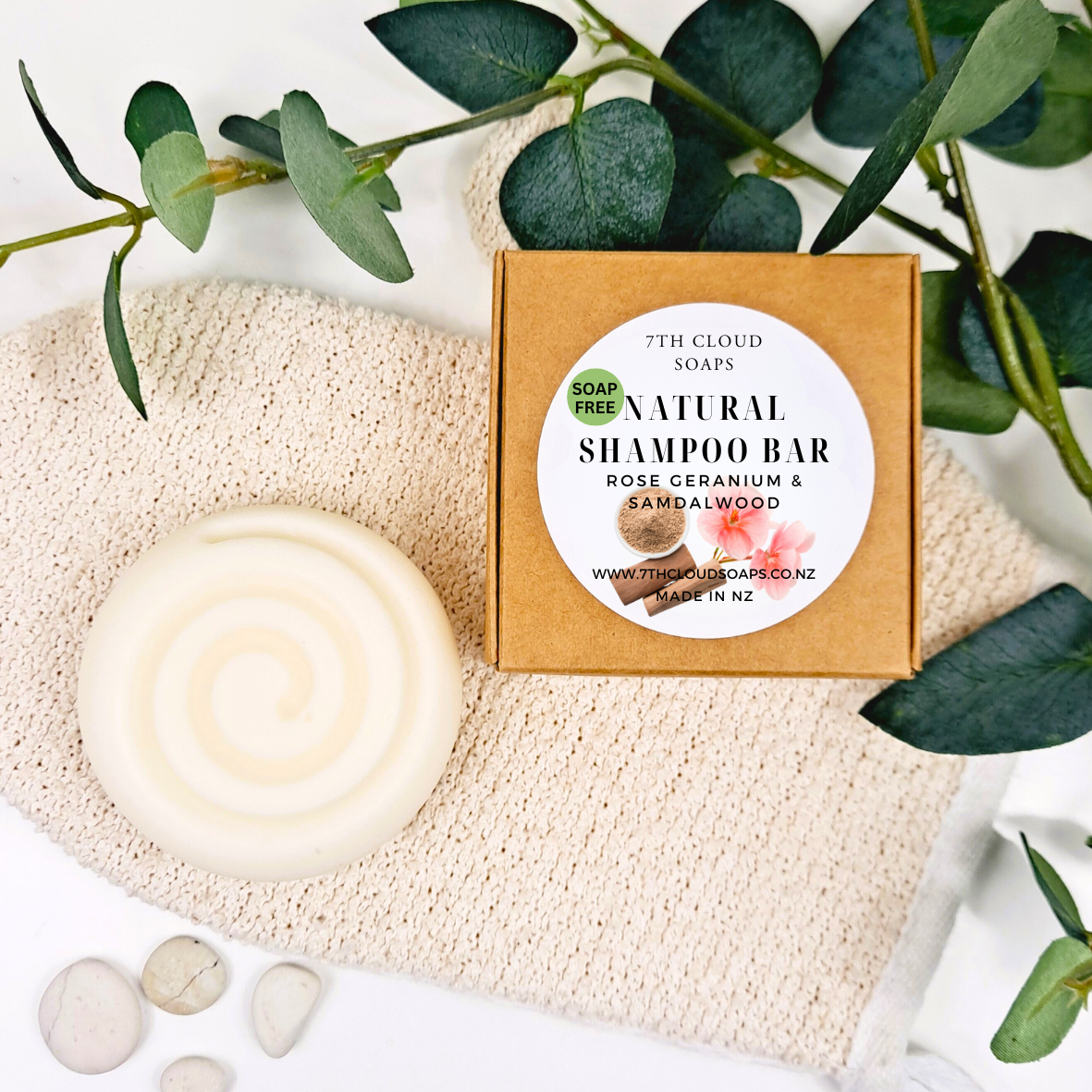 A natural shampoo bar with a solid white color, packaging box with product details, placed on a textured fabric with green leaves in the background.