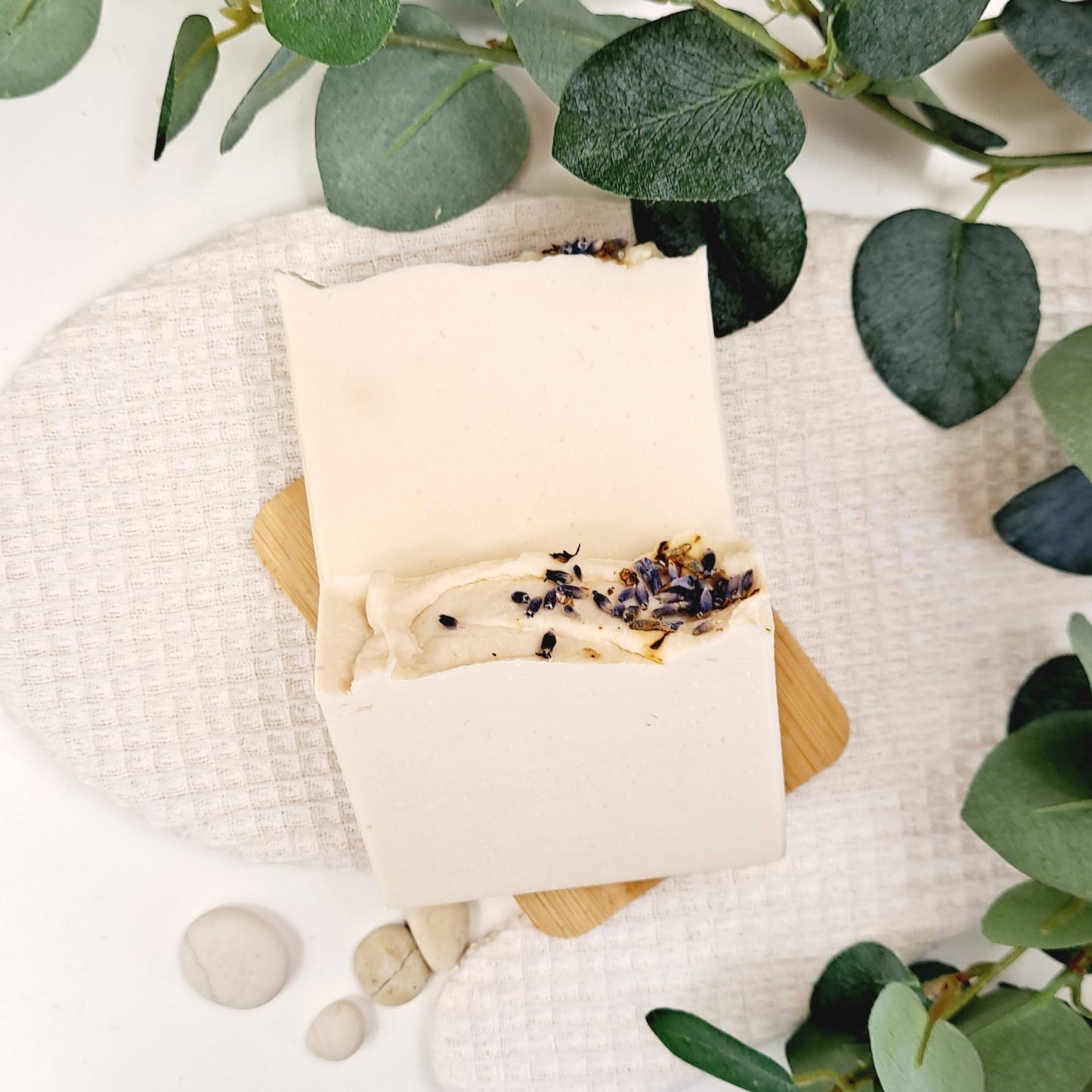 Bar of soap with lavender on a wooden soap dish, surrounded by green leaves and stones.