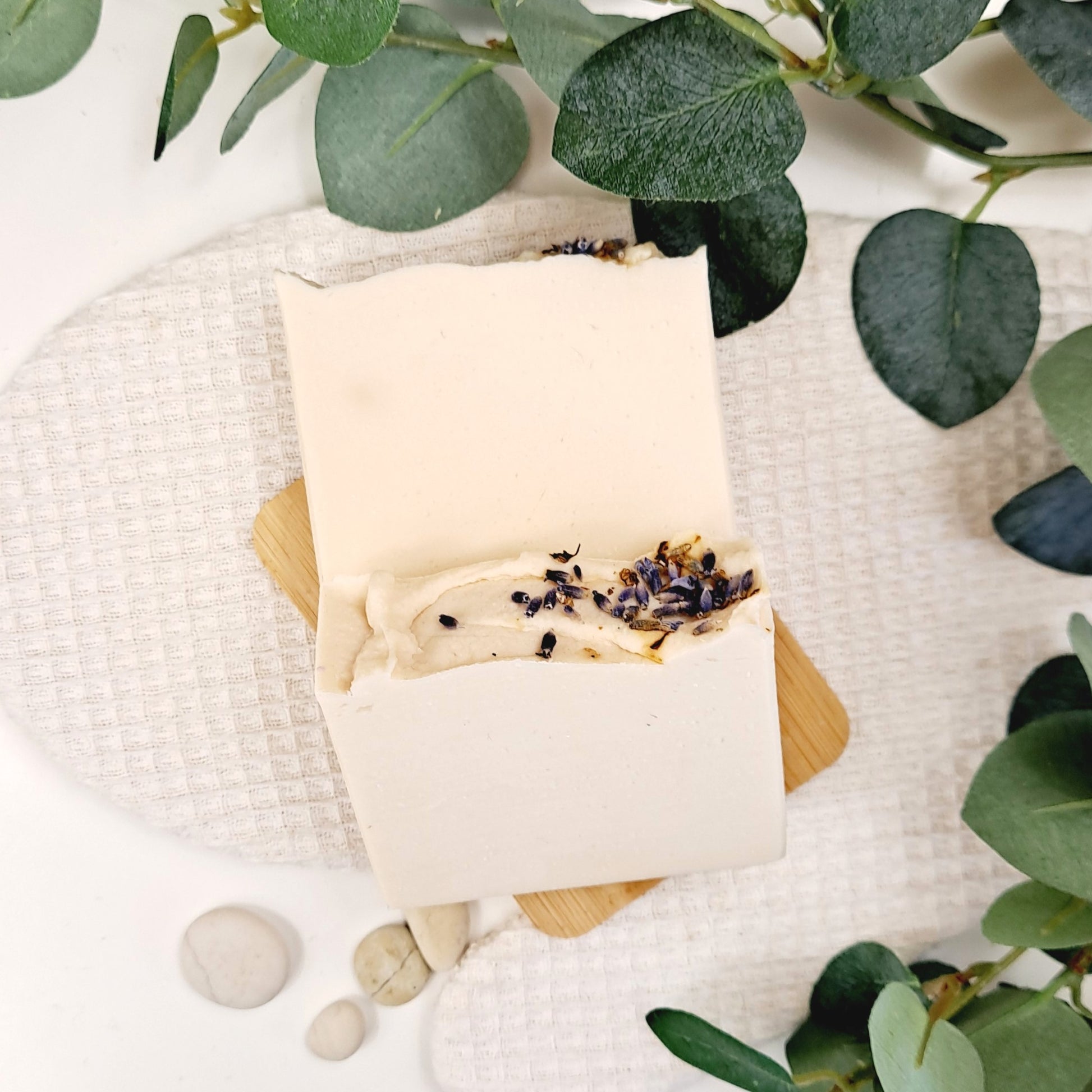 Bar of soap with lavender on a wooden soap dish, surrounded by green leaves and stones.