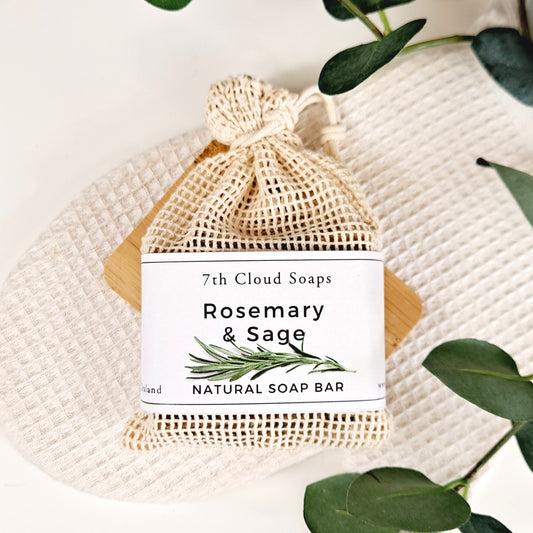 A natural bar of soap labeled 'Rosemary & Sage' from 7th Cloud Soaps, placed inside a beige netted bag, with green leaves in the background.
